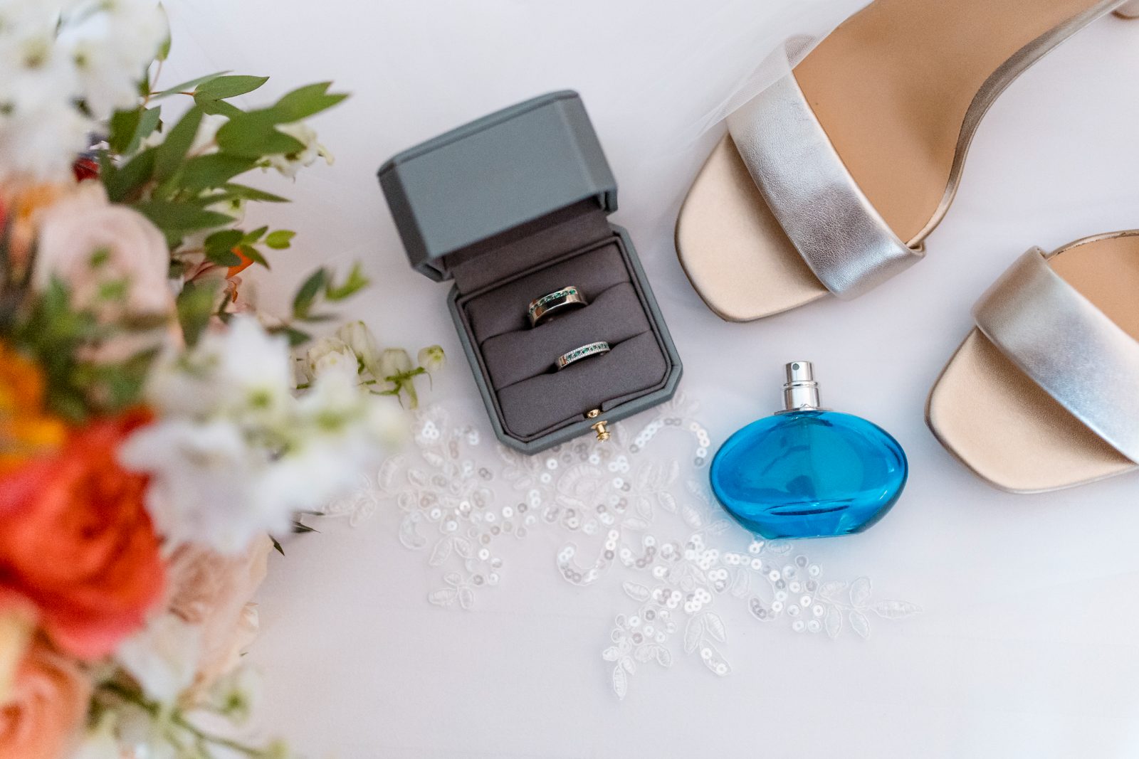 The scene shows wedding rings in a box, a pair of silver heels, perfume, and a floral arrangement, set against a white background
