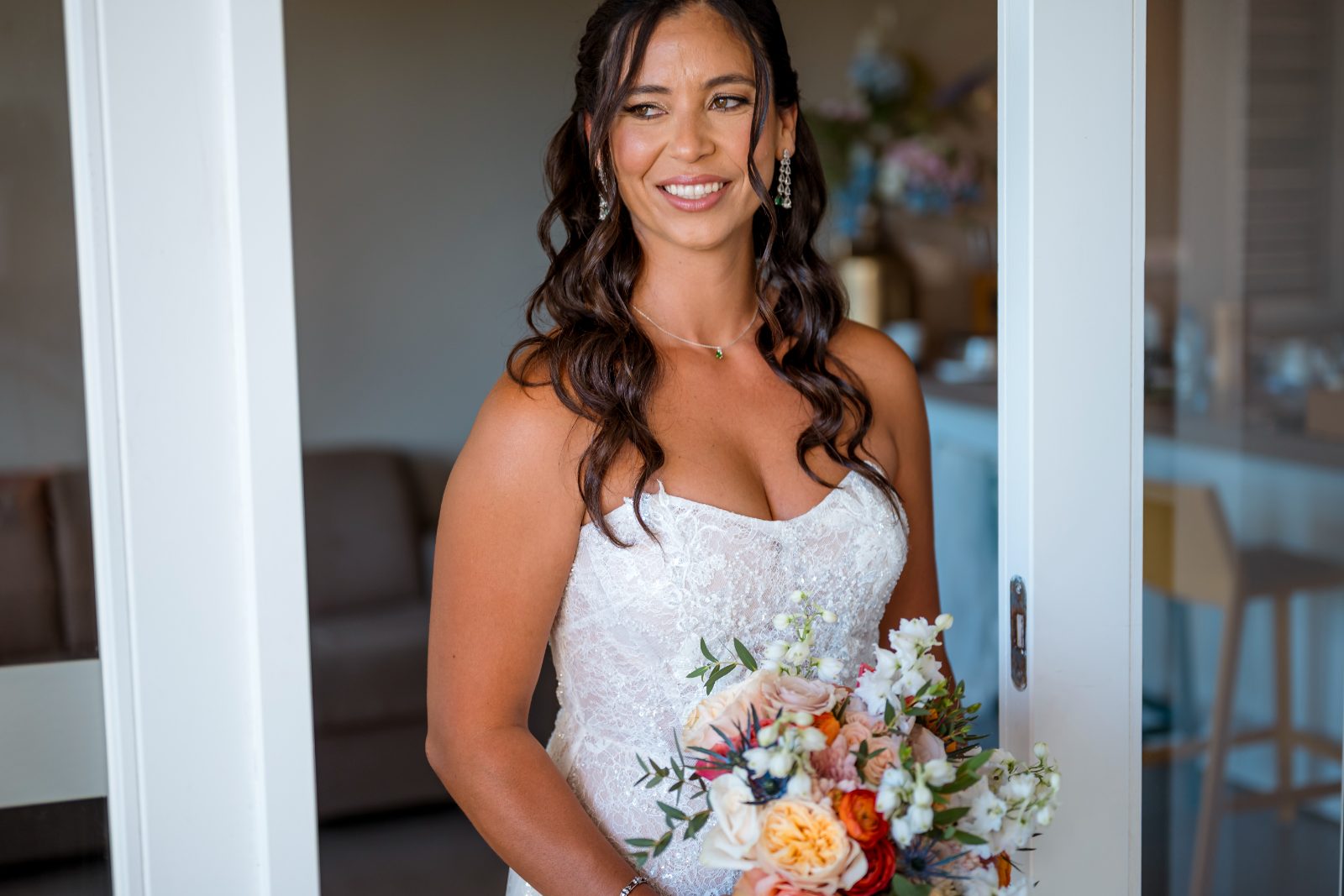Anika stands in a doorway holding a bouquet, wearing a wedding dress, at the Avila Beach Hotel with a soft interior background