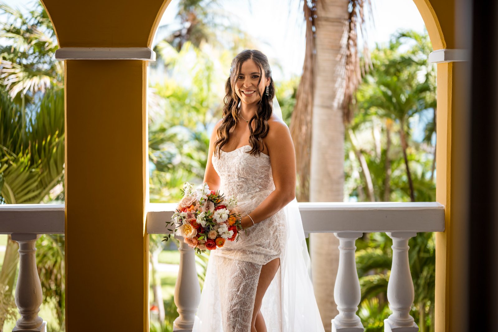 The bride stands on a balcony at Avila Beach Hotel, holding a bouquet of flowers amidst tropical greenery