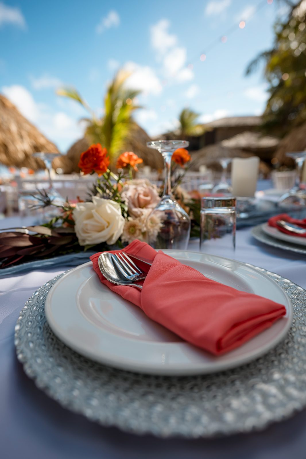 A wedding table is set at Avila Beach Hotel with a coral napkin, cutlery, and centerpieces featuring flowers, under a blue sky