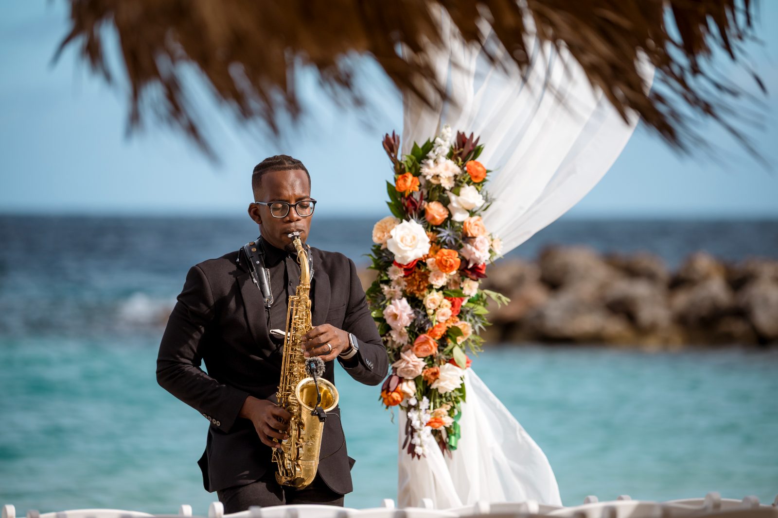 The wedding ceremony at Avila Beach Hotel features a musician playing saxophone near a floral arch and the ocean in the background
