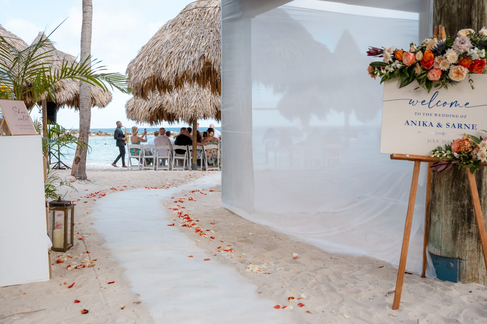 The wedding ceremony for Anika and Sarne is set at Avila Beach Hotel, with guests seated under thatched roofs beside a sandy walkway