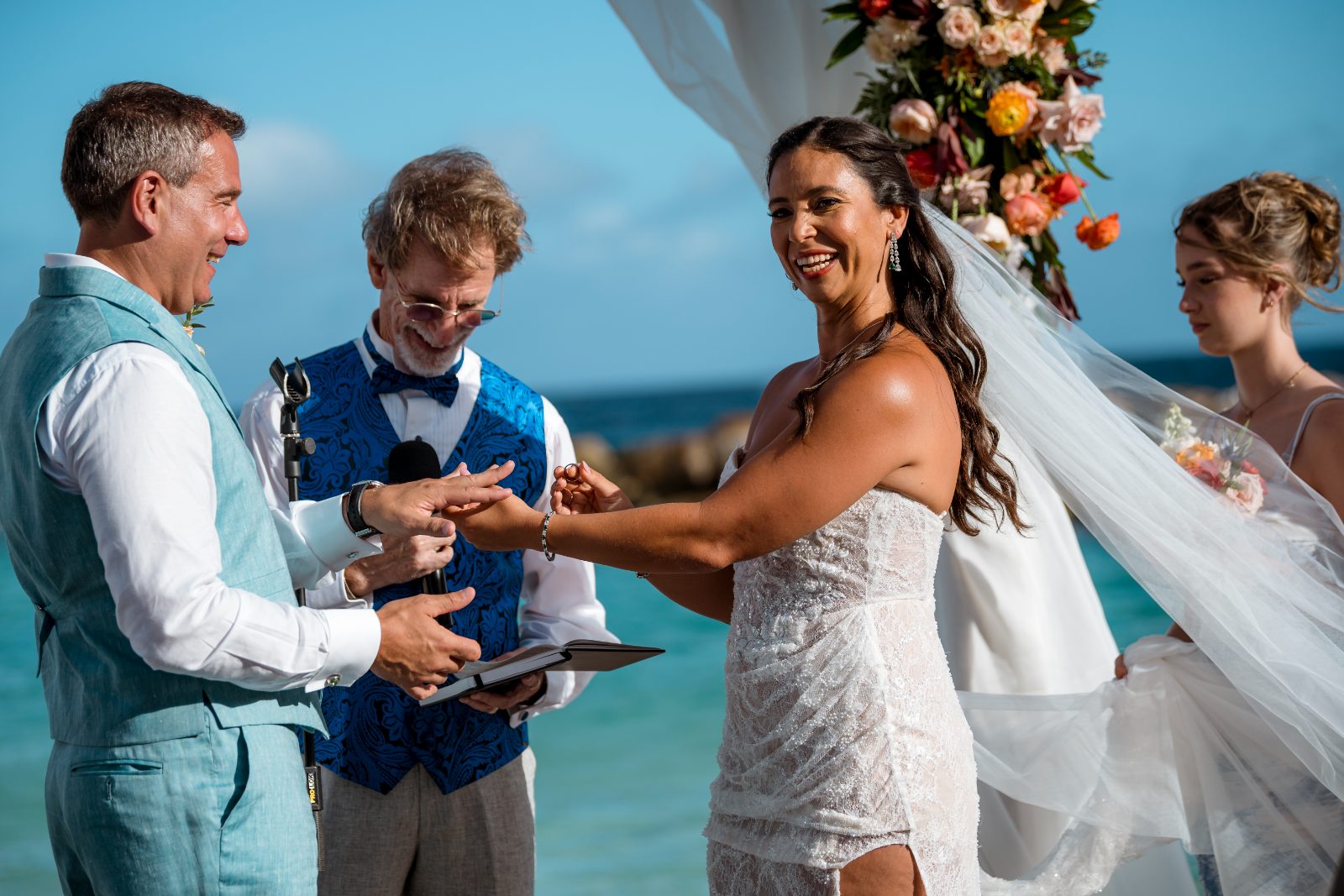 Anika and Sarne are exchanging vows at their wedding ceremony on the beach, with a celebrant and a bridesmaid present at Avila Beach Hotel