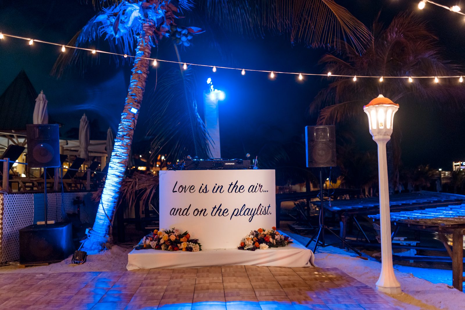 Anika and Sarne stand in front of a decorated backdrop with a DJ setup at night, surrounded by lights and flowers at Avila Beach Hotel