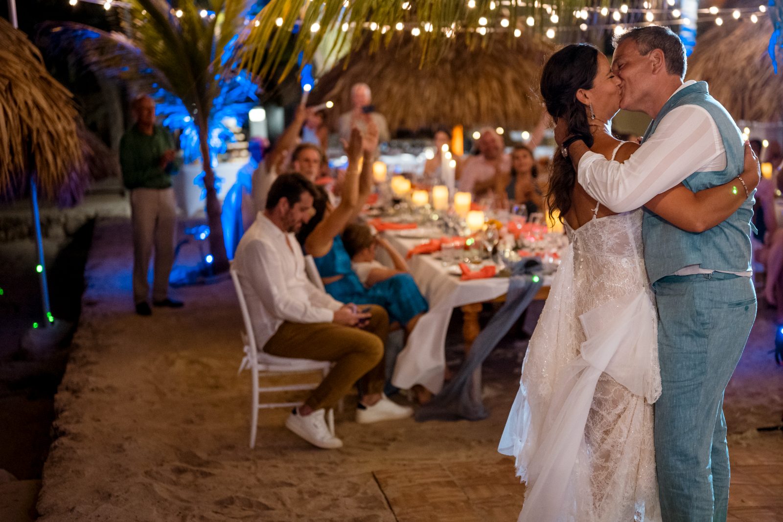 Anika and Sarne are kissing while surrounded by guests at their wedding reception on the beach at Avila Beach Hotel, with decorations and candles in the background