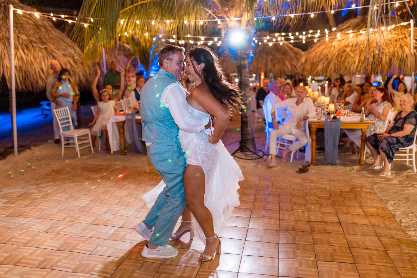Anika and Sarne are dancing together at their wedding reception on the beach, with guests seated at tables nearby under string lights