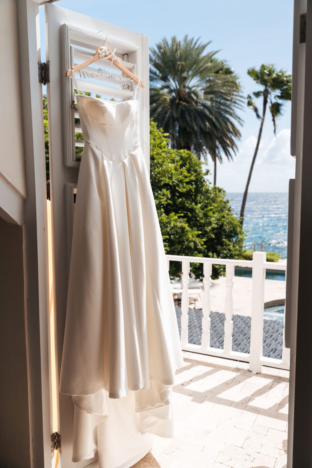 A wedding dress hangs on a hanger in a doorway, with palm trees and the ocean visible outside at the Avila Beach Hotel, Curaçao