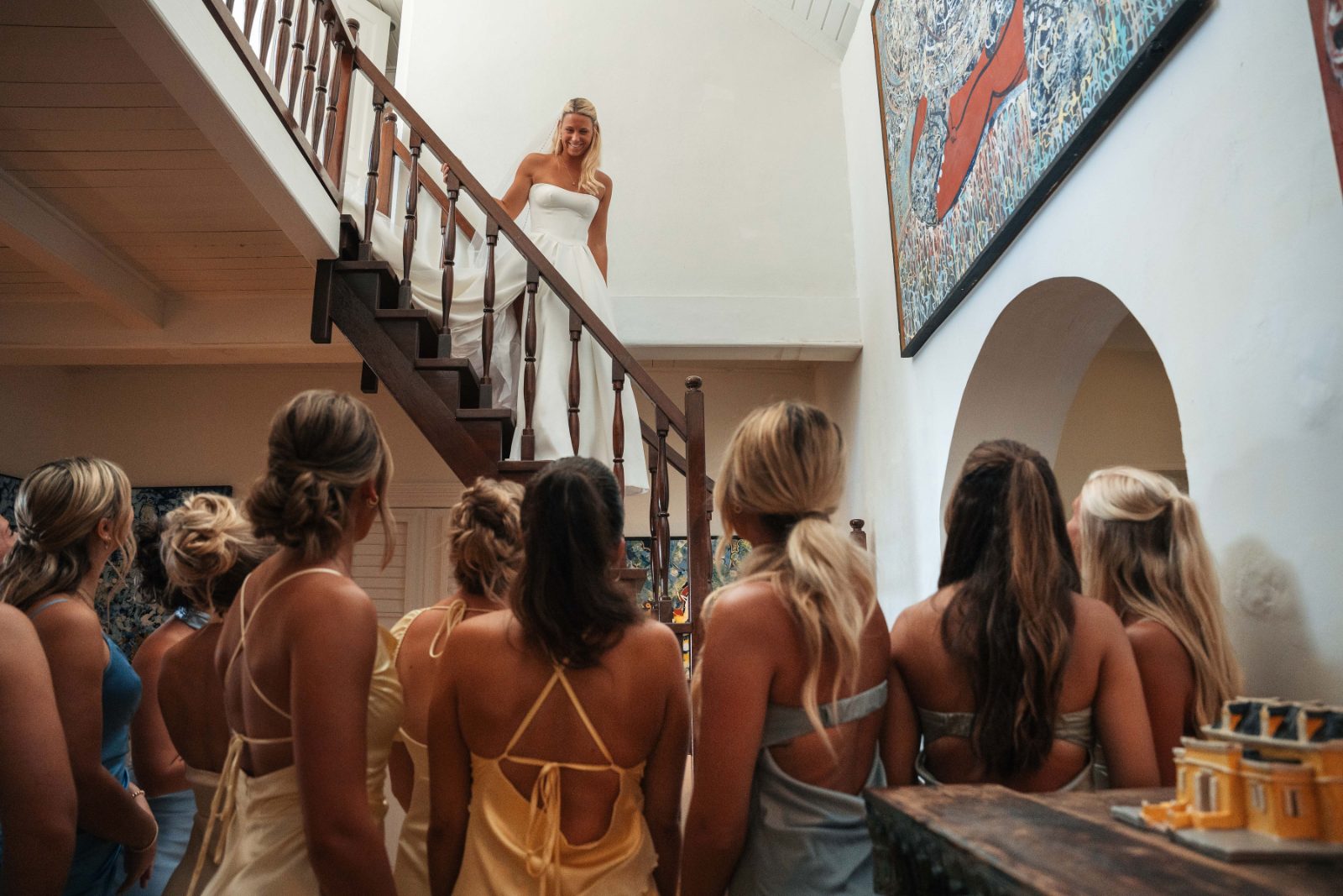 Cailey stands on the stairs in her wedding dress at Avila Beach Hotel, while her bridesmaids, in pastel gowns, look up at her