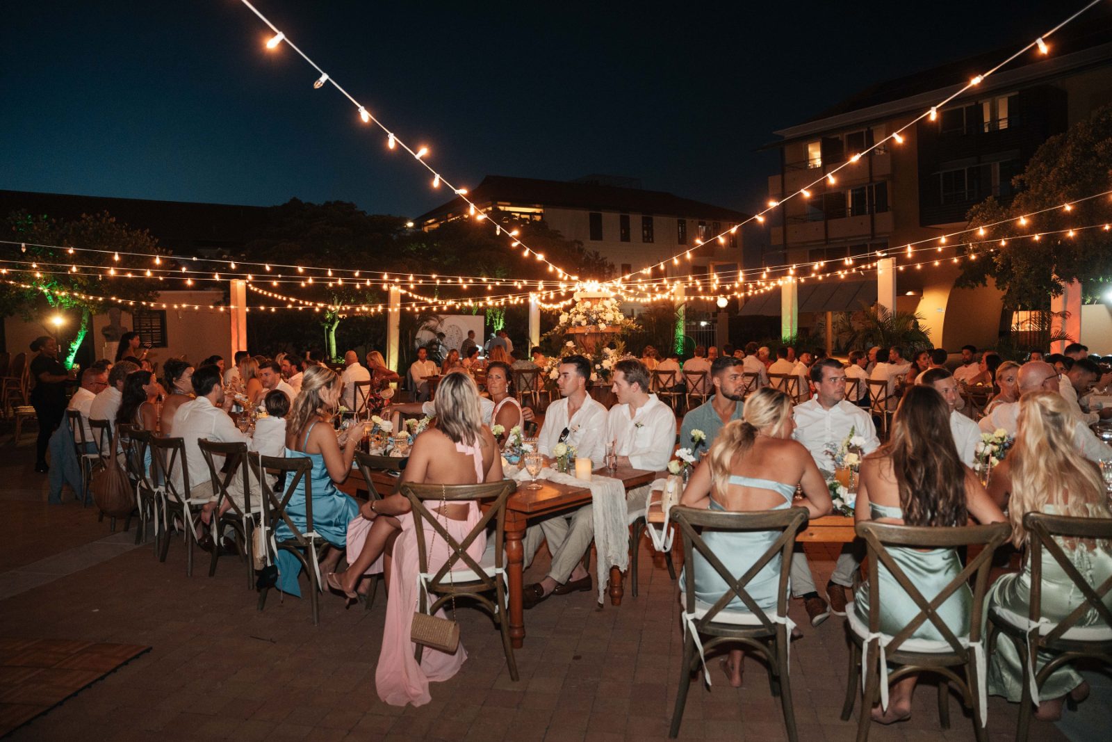 Cailey and Noel's wedding reception is set at Avila Beach Hotel in Curaçao, with guests seated at tables illuminated by string lights at night