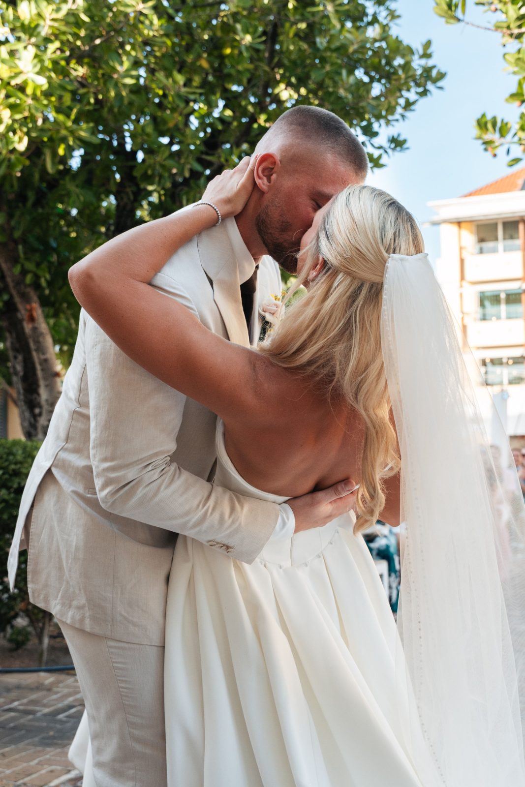 Cailey and Noel are kissing at their wedding ceremony at Avila Beach Hotel in Curaçao, surrounded by greenery and a visible building in the background