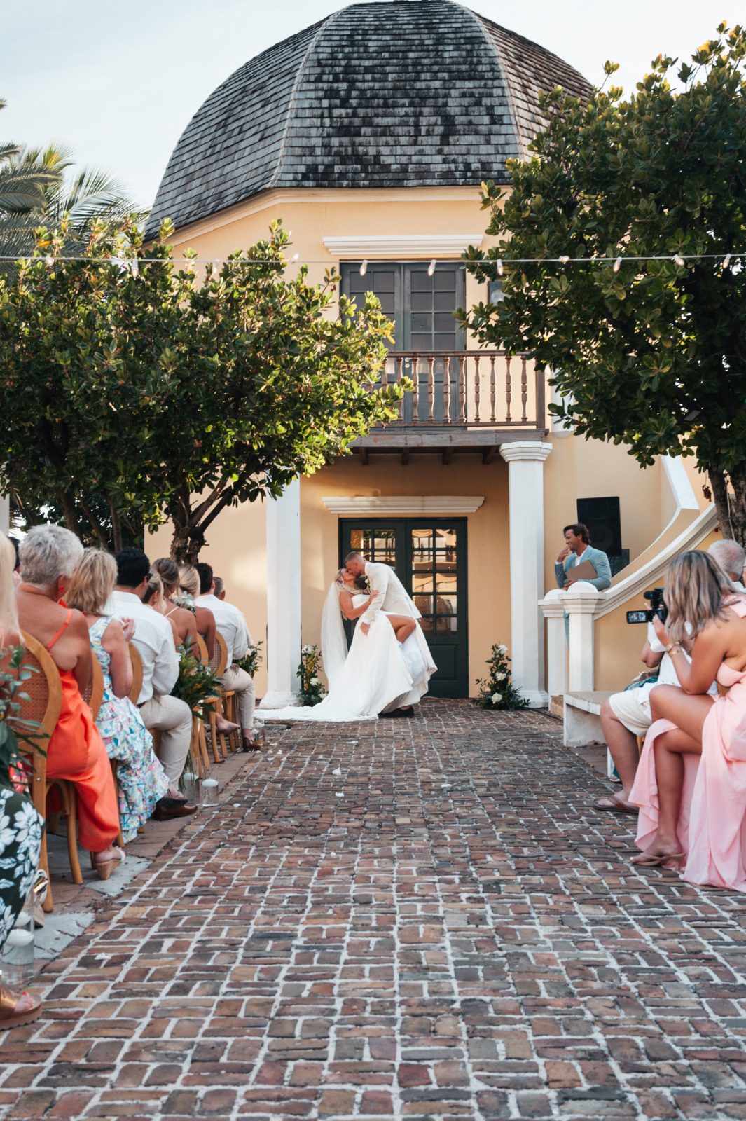 Cailey and Noel are exchanging vows outdoors at the Avila Beach Hotel in Curaçao, surrounded by seated guests and greenery