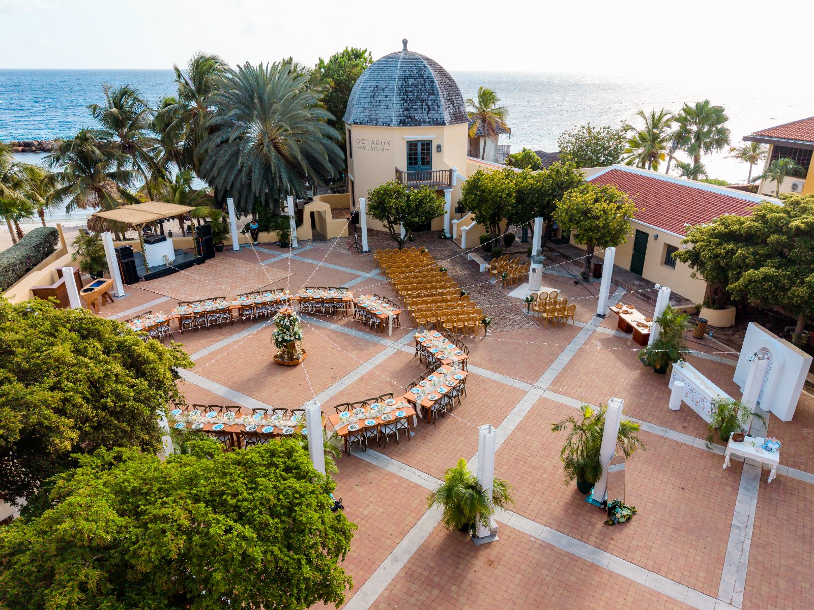 Cailey and Noel's wedding ceremony takes place at the Avila Beach Hotel in Curaçao, featuring a circular arrangement of chairs surrounded by trees and ocean views