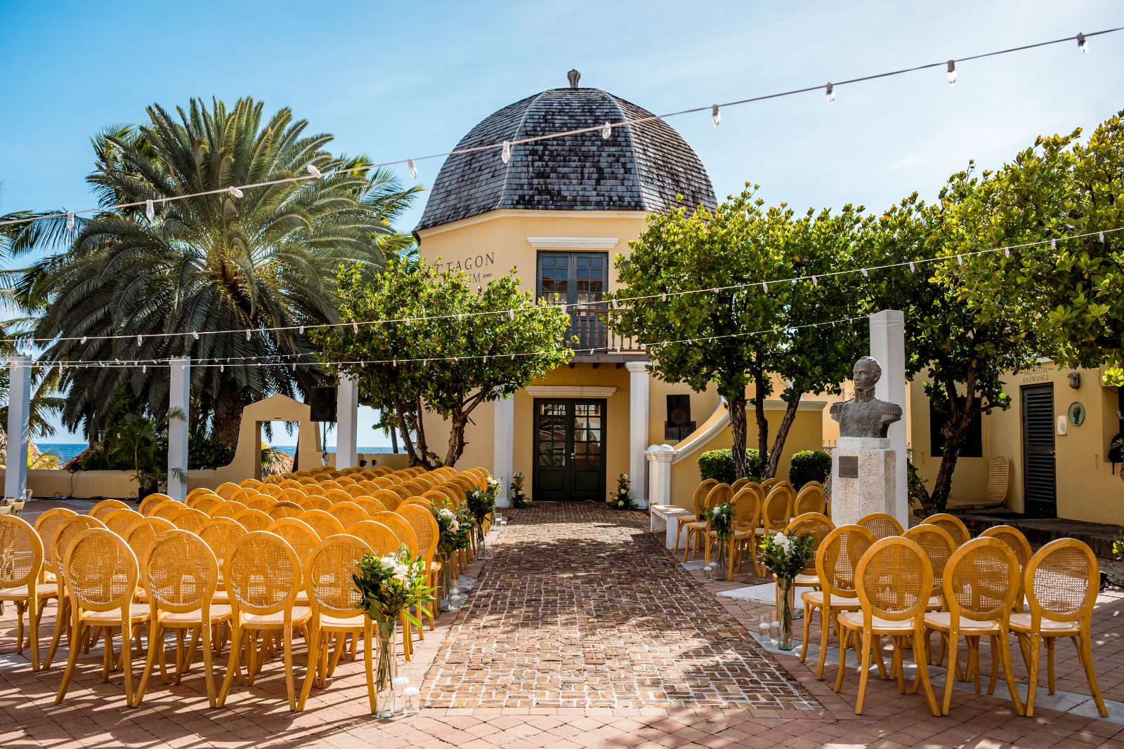 Cailey and Noel are getting married at Avila Beach Hotel in Curaçao, surrounded by empty chairs arranged for the ceremony near palm trees and greenery
