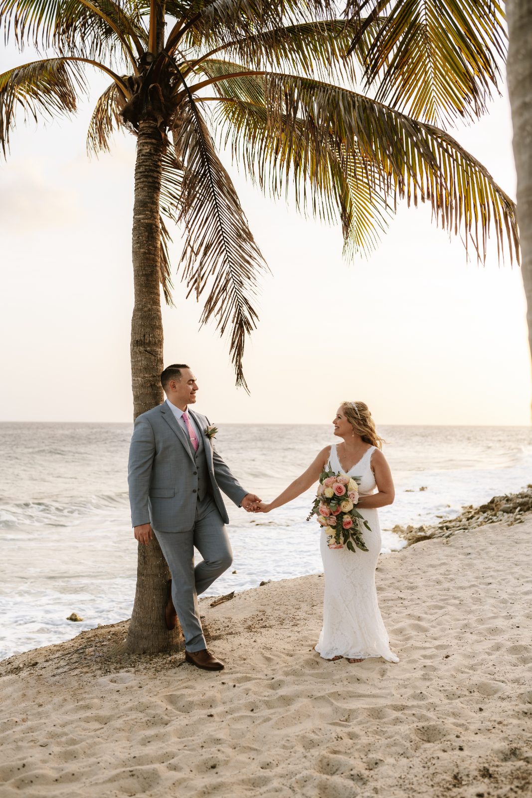 Christine and Jesse hold hands by the beach at a private villa in Curaçao, with palm trees and the ocean in the background