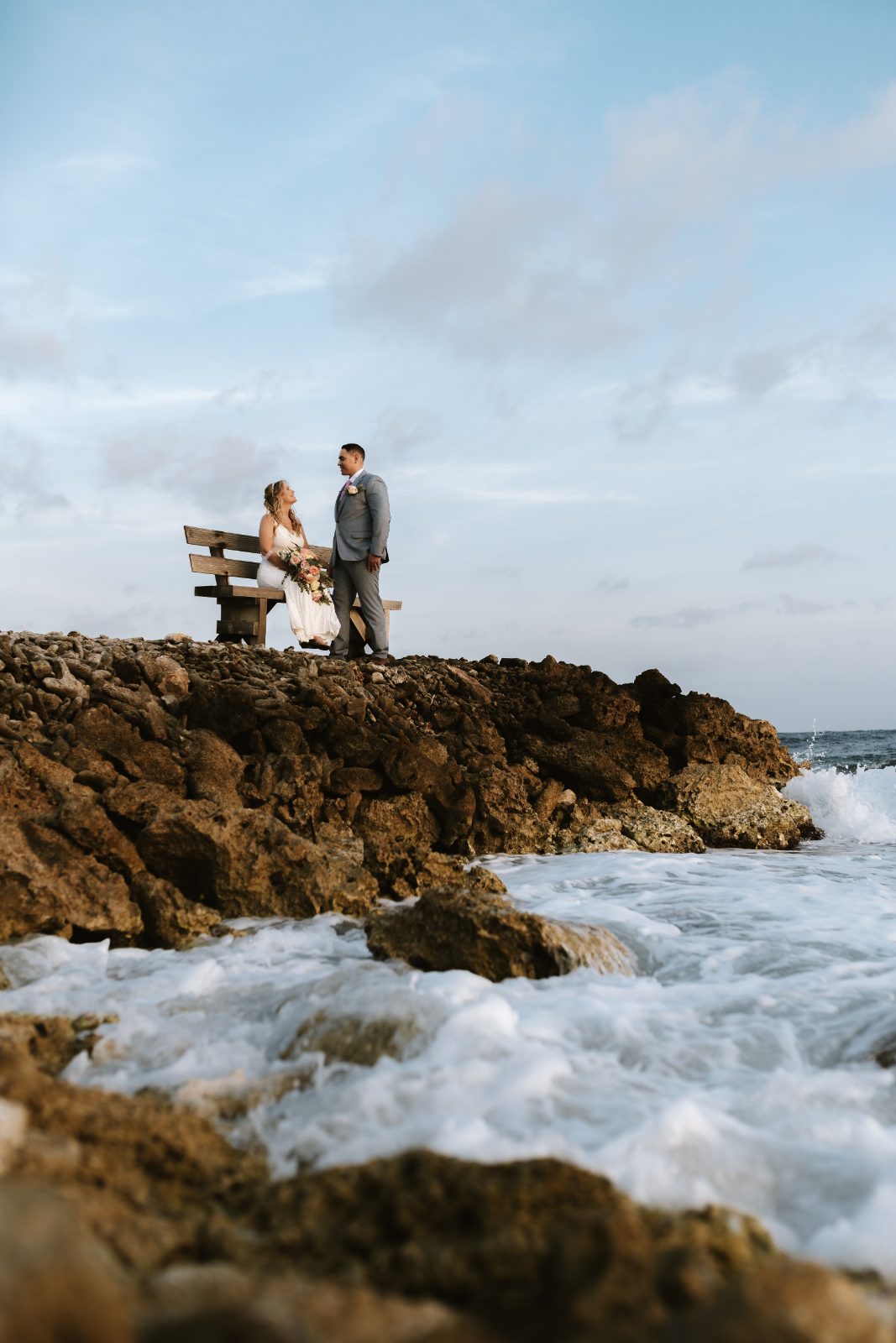Christine and Jesse stand on a rocky shore near a bench, with waves crashing against the rocks in Curaçao
