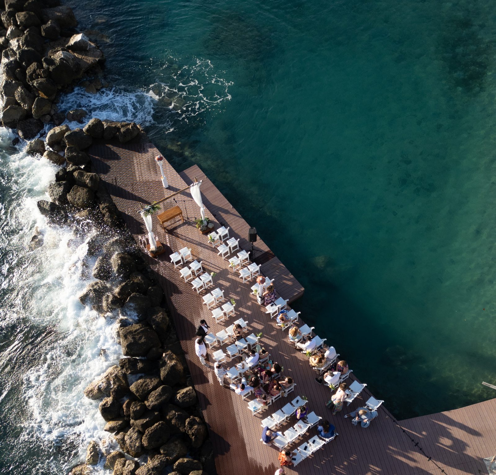 Erica and Derek's wedding ceremony takes place on a wooden dock by the ocean at Avila Beach Hotel in Curaçao, with guests seated on white chairs