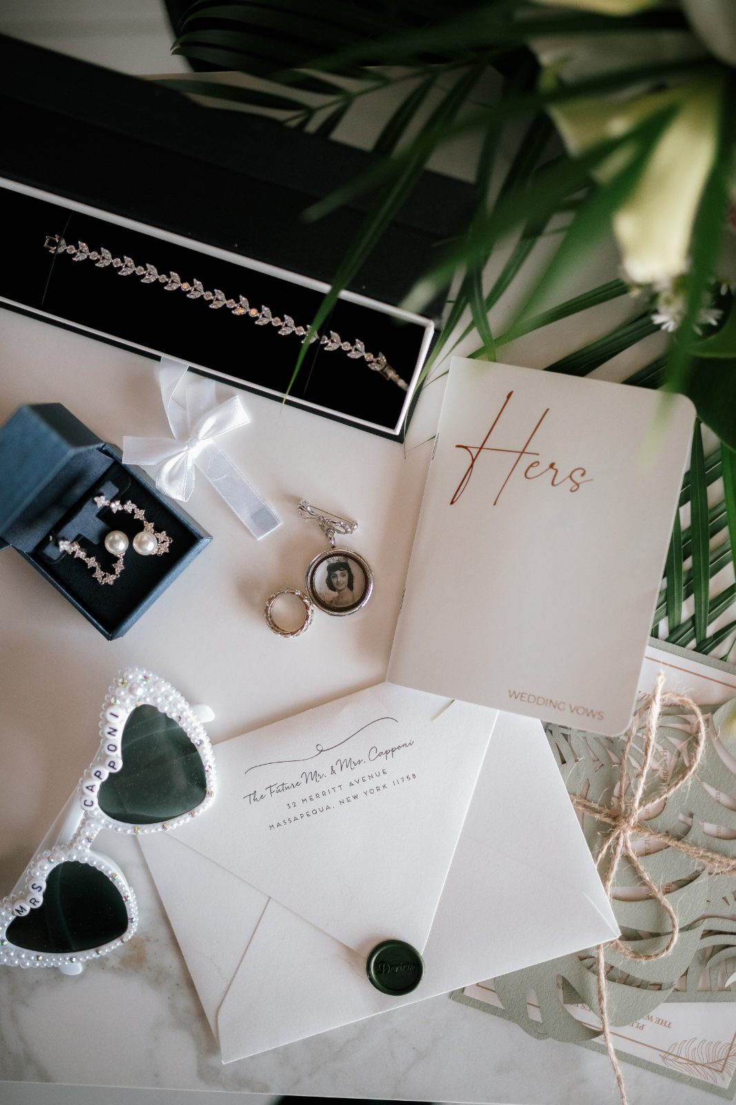 The wedding details are displayed on a table, including jewelry, vows, and themed accessories, set against a backdrop of greenery