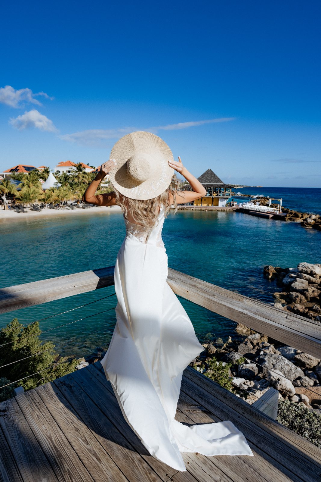Erica stands by the water at Avila Beach Hotel in Curaçao, wearing a wedding dress and a large sun hat, looking out at the ocean