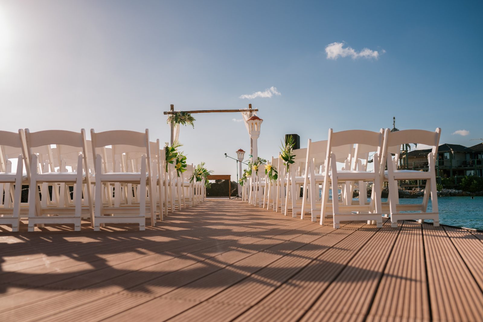 Erica and Derek's wedding ceremony takes place at Avila Beach Hotel in Curaçao, with white chairs arranged on a wooden dock overlooking the water