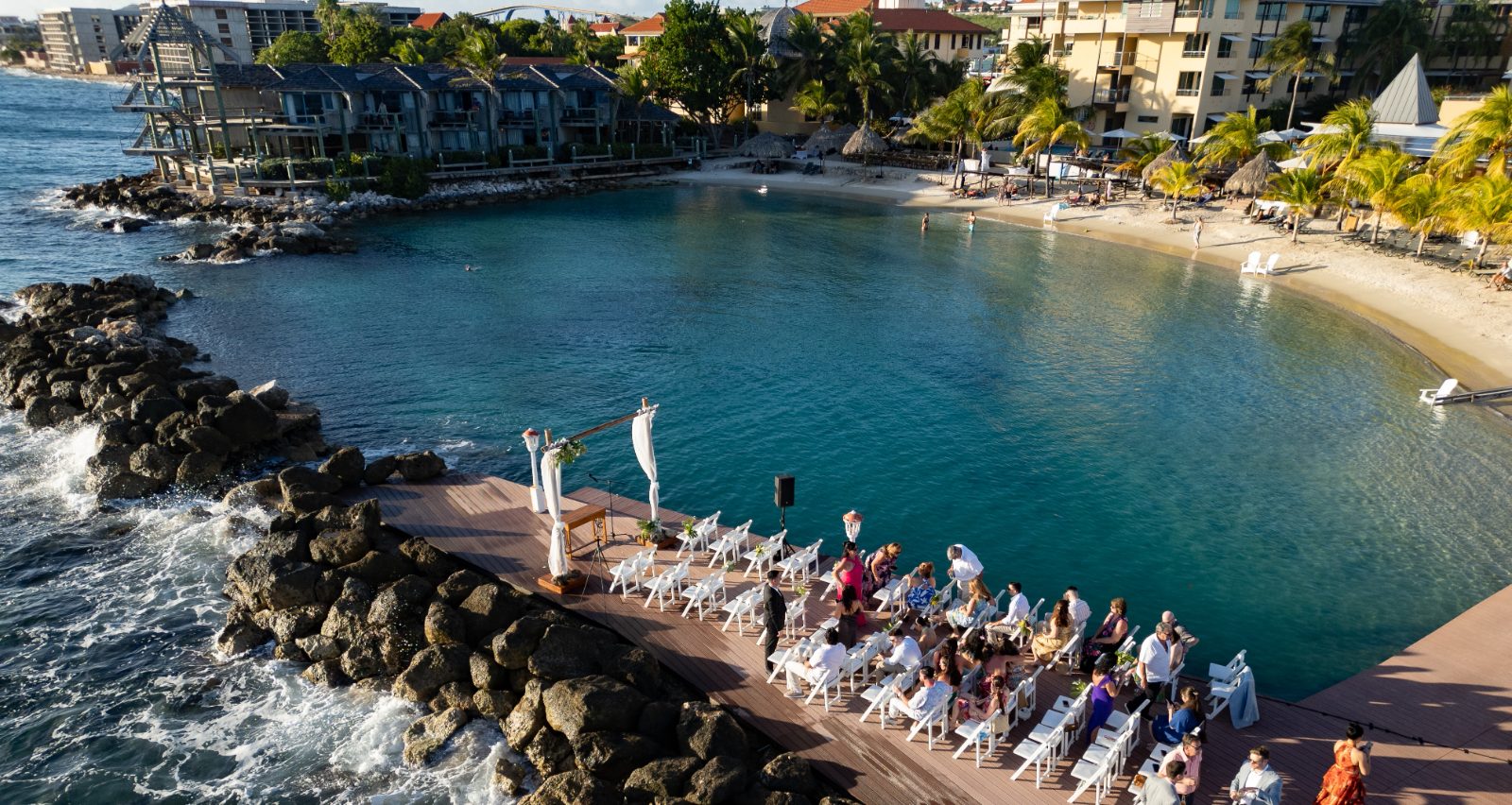 Erica and Derek are getting married on a wooden dock overlooking a beach and calm sea at Avila Beach Hotel in Curaçao, with guests seated