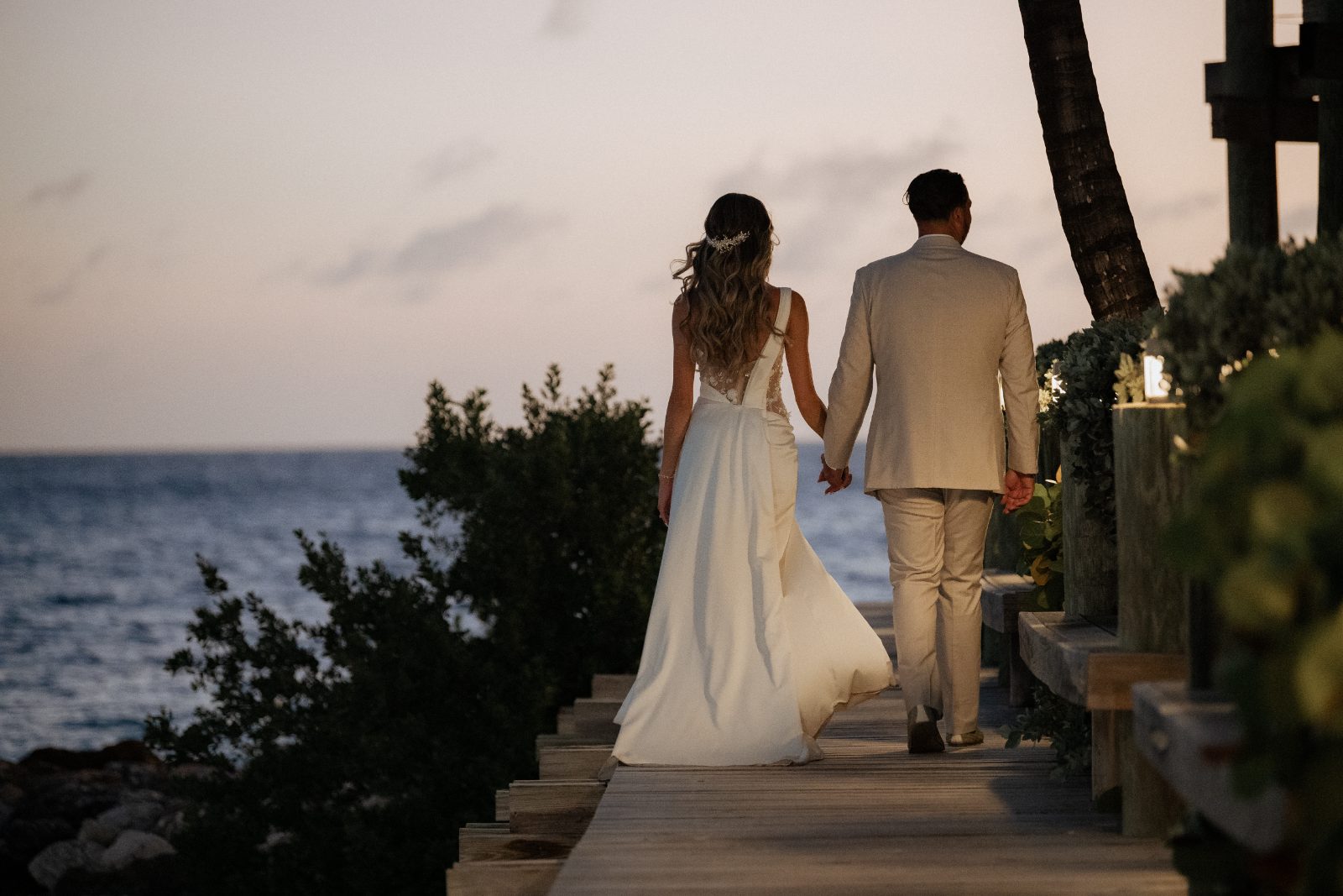 Erica and Derek walk hand in hand along a wooden path by the ocean at Avila Beach Hotel in Curaçao during sunset