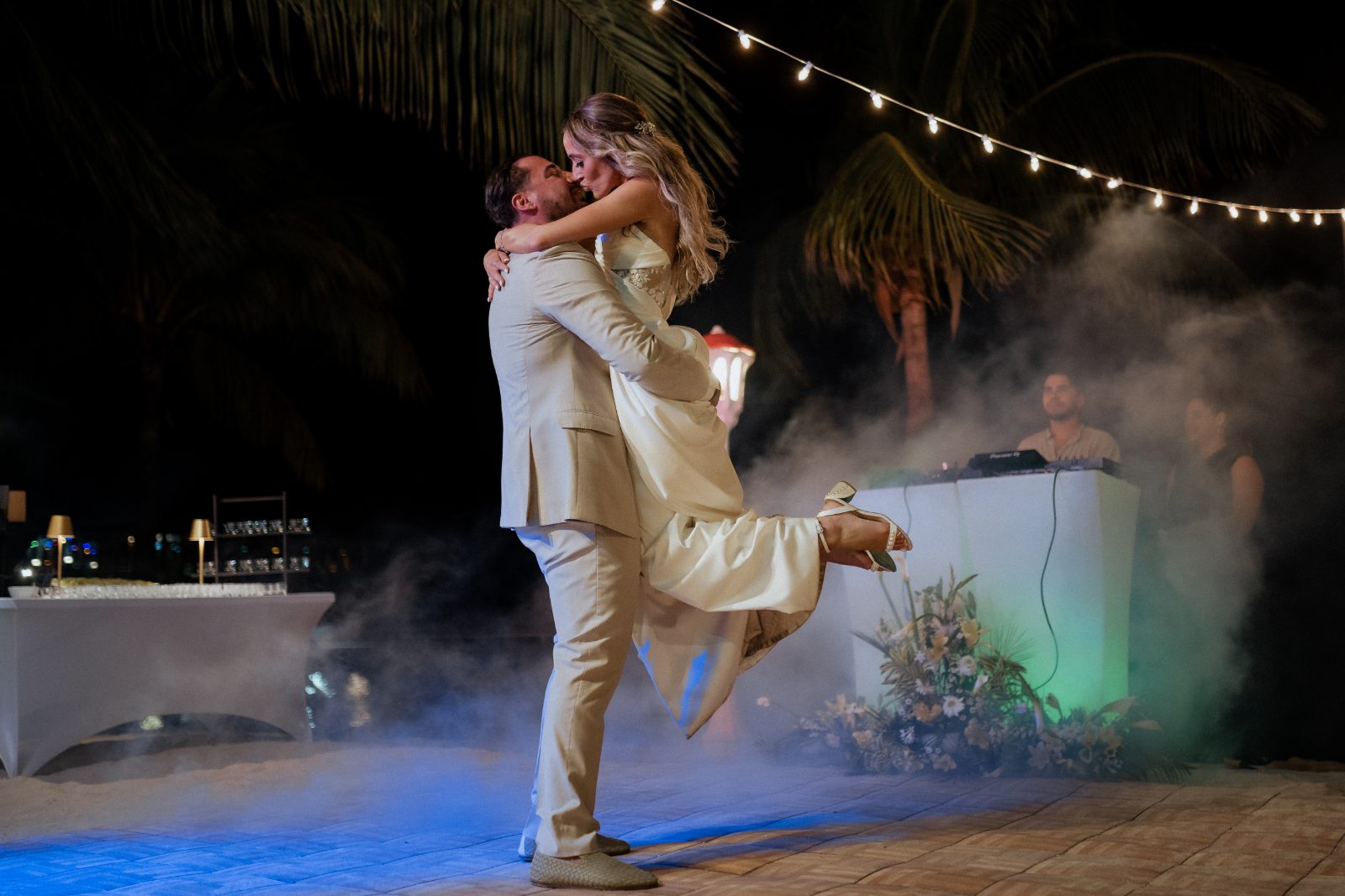Erica and Derek celebrate their wedding at Avila Beach Hotel, Curaçao, as Derek lifts Erica amid a festive setting with lights and smoke