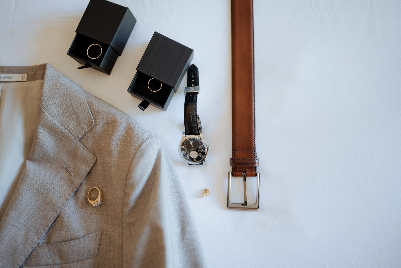 Flatlay attire and accessories, including a suit, watch, rings in boxes, and a belt, are arranged on a flat surface at a beach resort