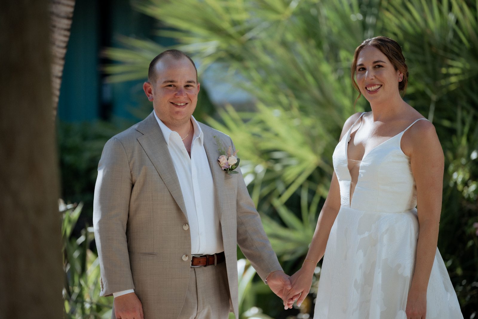 Frannie and Dustin stand together, holding hands, smiling in front of palm trees at the Curaçao Marriott Beach Resort during their first look