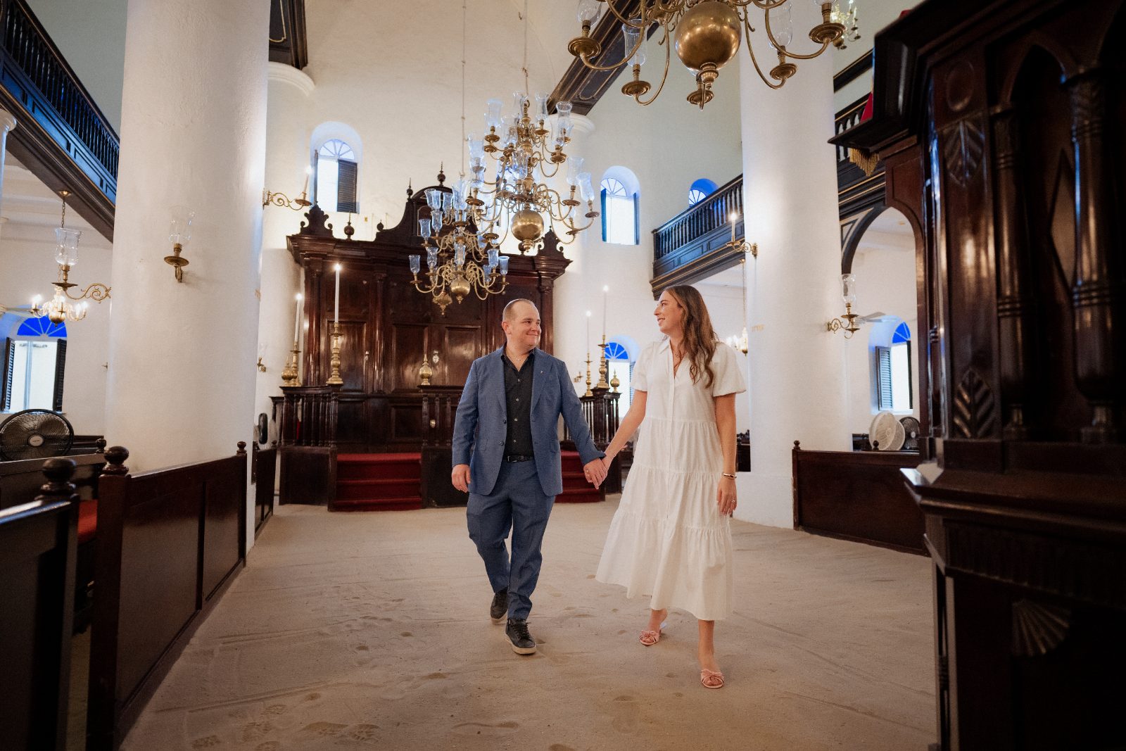 Frannie and Dustin walk hand in hand inside Mikvé Israel-Emanuel Synagogue, surrounded by ornate chandeliers and wooden details