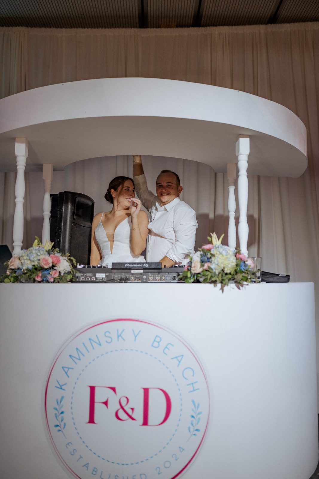 Frannie and Dustin stand behind a DJ booth at their wedding reception, decorated with flowers, at the Curaçao Marriott Beach Resort