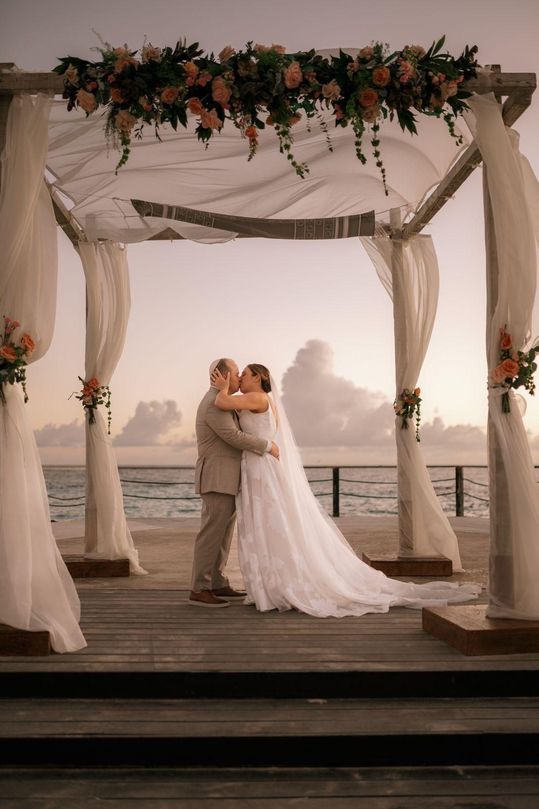 Frannie and Dustin kiss under a floral-draped canopy on the beach at Curaçao Marriott Beach Resort during their wedding ceremony