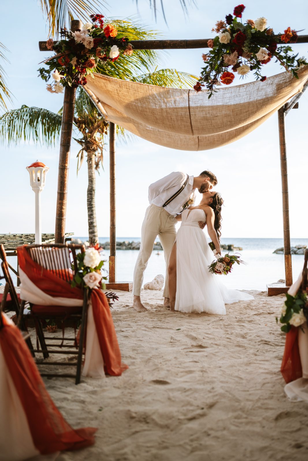 Gaëlle and Jeroen kiss during their wedding ceremony on the beach at Avila Beach Hotel in Curaçao, with decorated chairs in the foreground