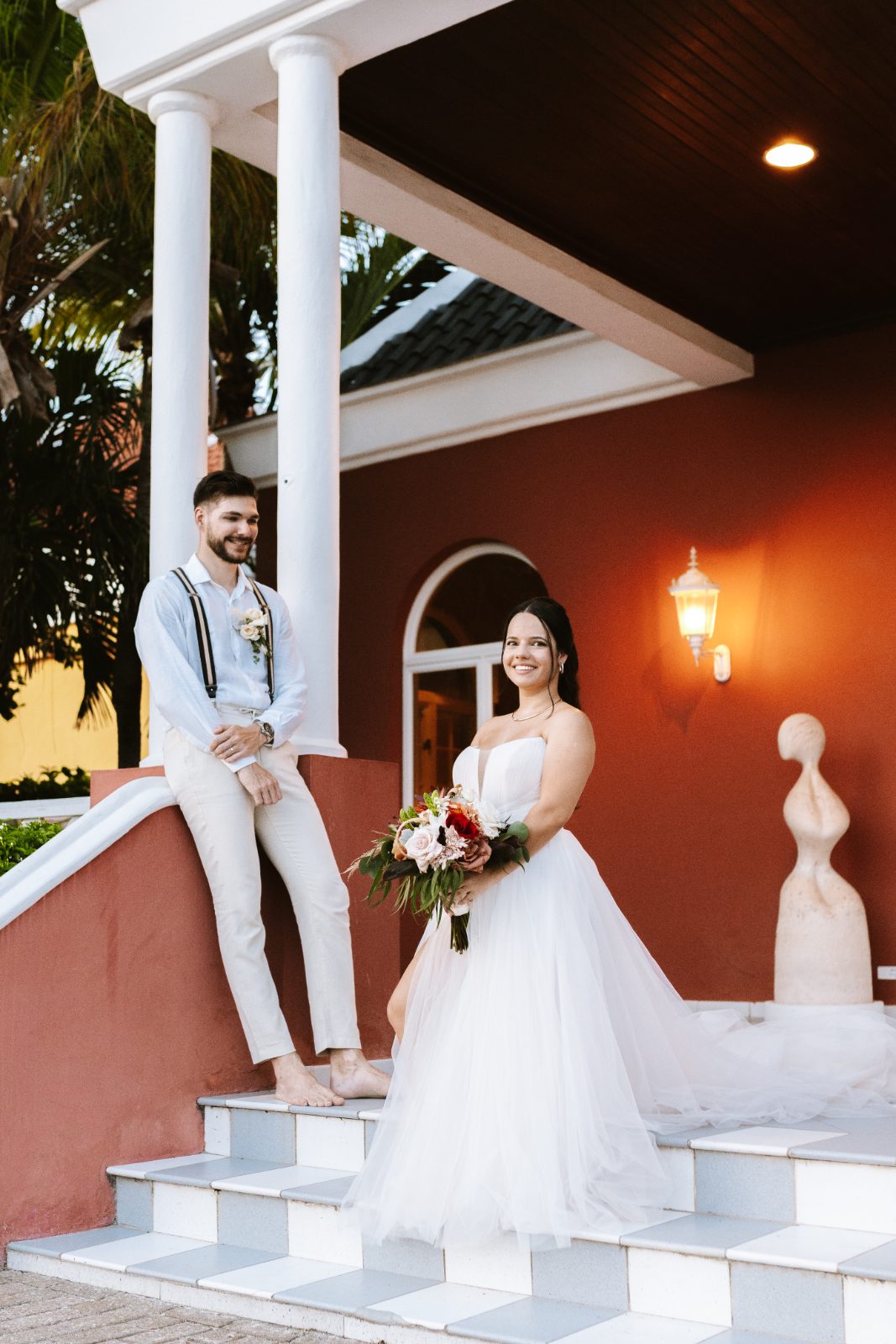 Gaëlle stands in her wedding dress holding a bouquet, while Jeroen poses beside her on the steps of the Avila Beach Hotel in Curaçao