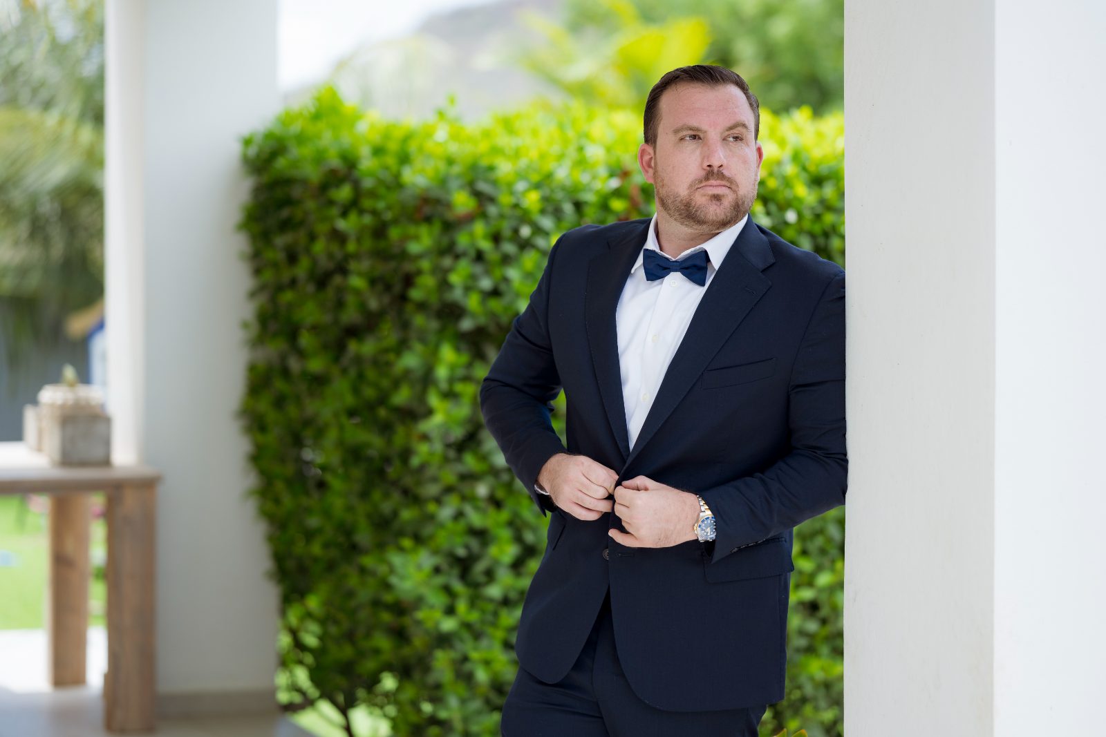 Kevin adjusts his tuxedo while standing beside a white column in a private garden in Curaçao, with greenery in the background