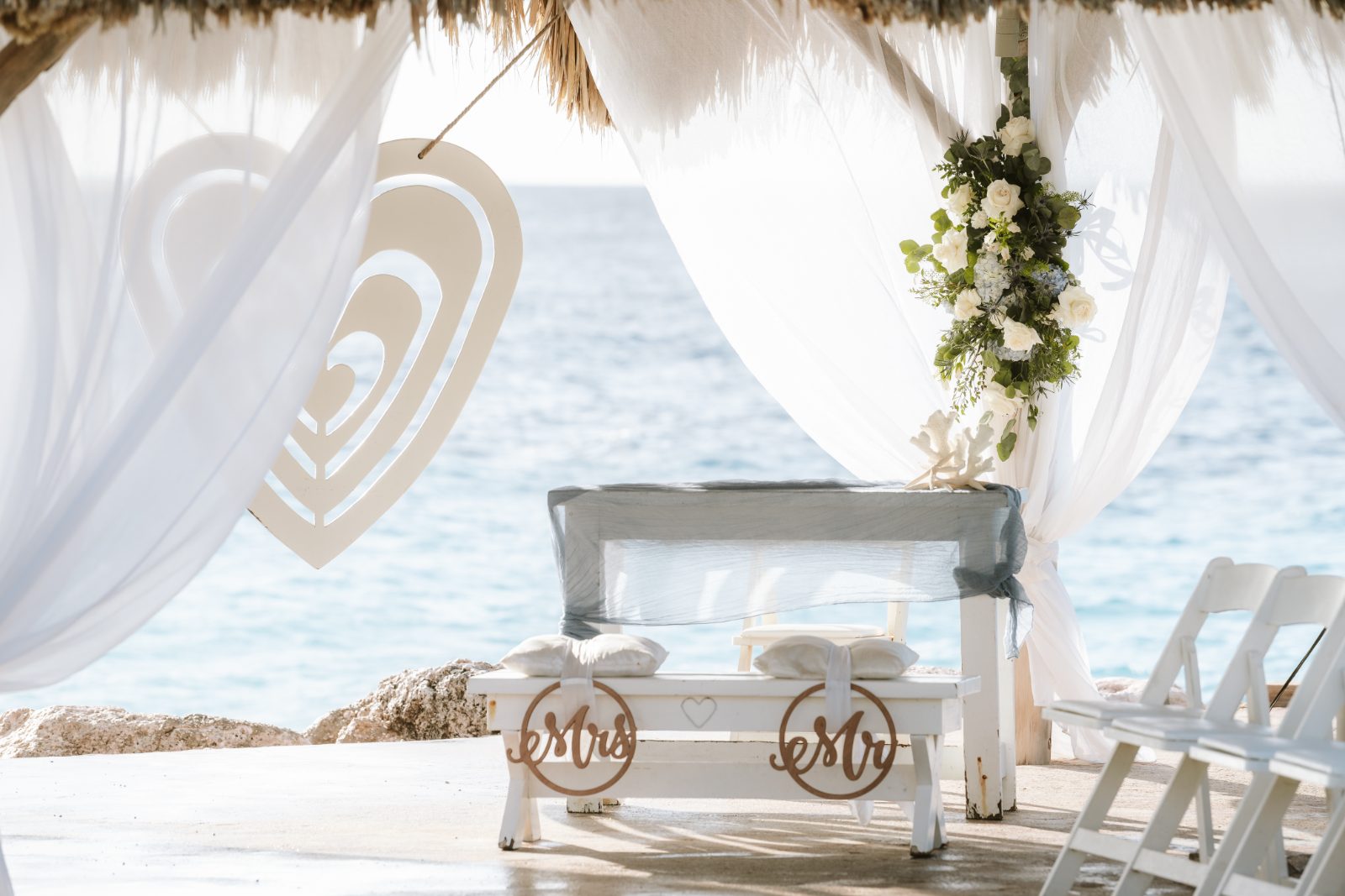 Jennifer and Weston are in a wedding venue at Karakter, Curaçao, with a decorated altar by the ocean and white draped fabric