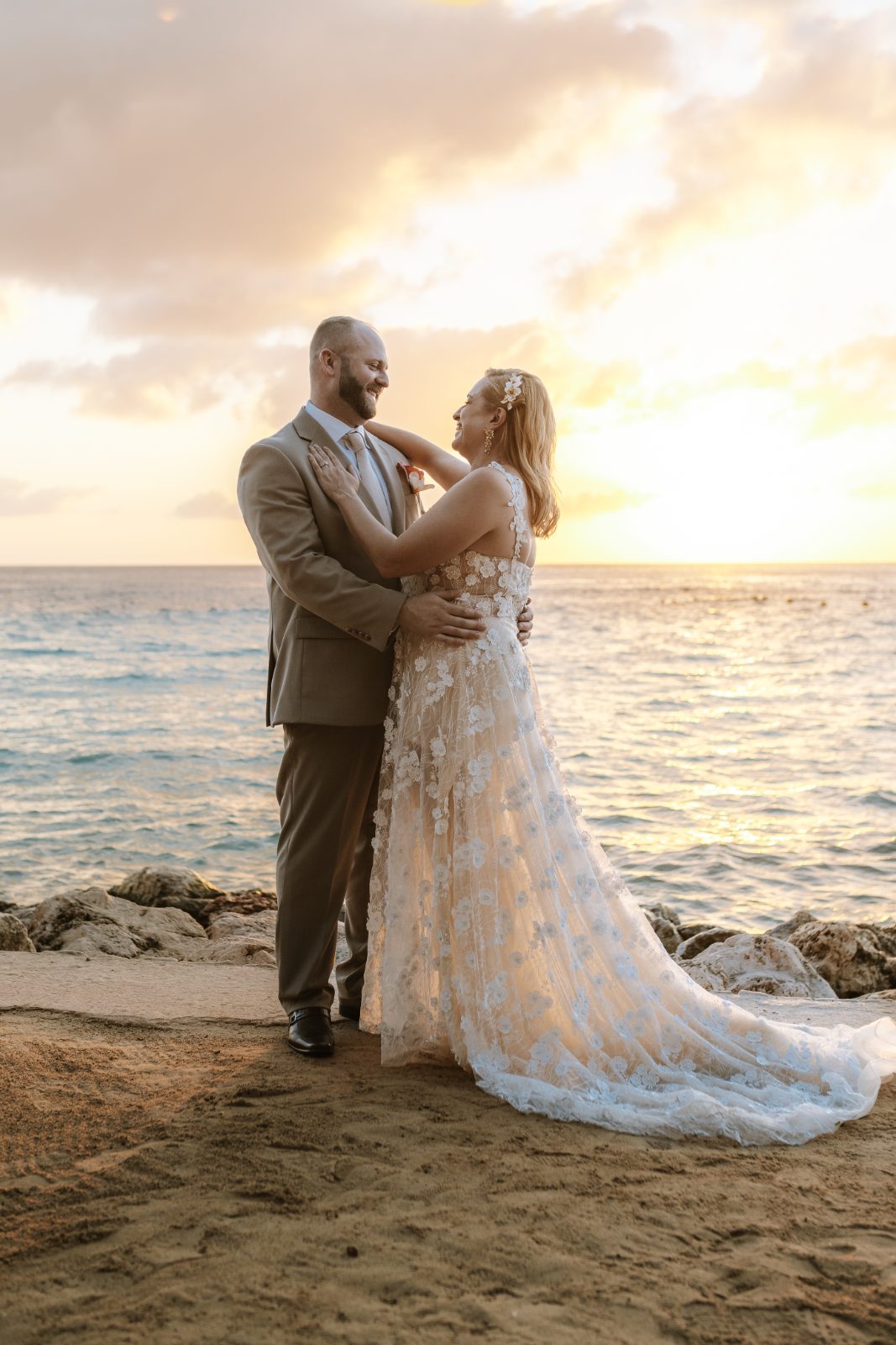 Jennifer and Weston stand together on a beach at dusk, overlooking the ocean with a sunset in the background