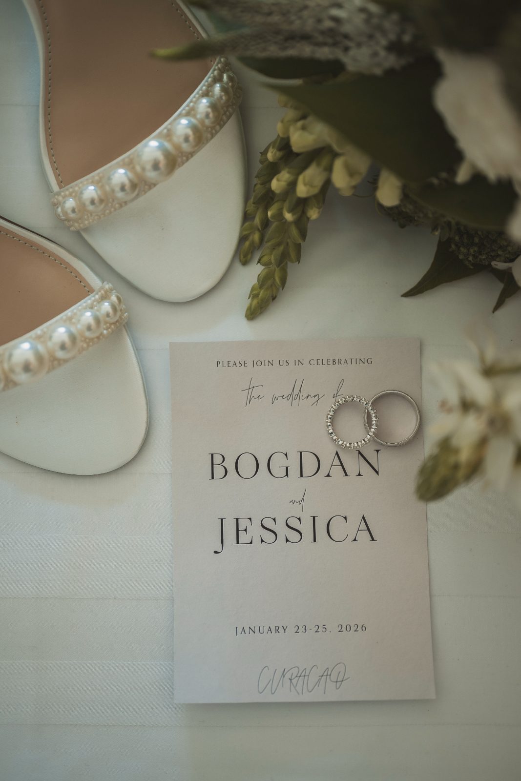 Jessica and Bogdan's wedding invitation lies on a flat surface next to white wedding shoes and floral arrangements at Avila Beach Hotel, Curaçao