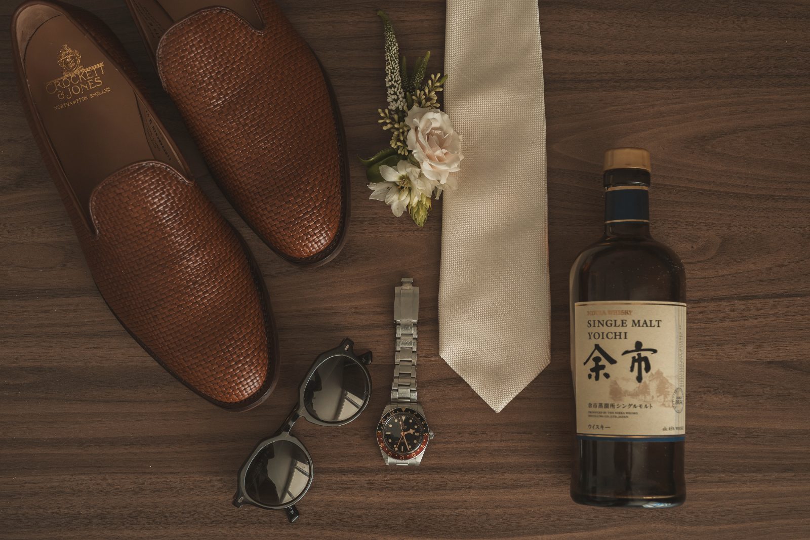 The wedding items, including shoes, a watch, sunglasses, a tie, a flower bouquet, and a whiskey bottle, are arranged on a wooden surface