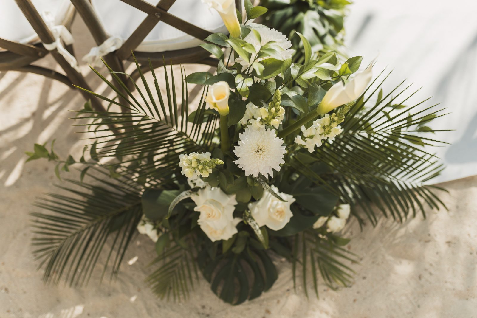 Jessica and Bogdan stand under a floral arch at Avila Beach Hotel in Curaçao, surrounded by white flowers and greenery on the sandy ground