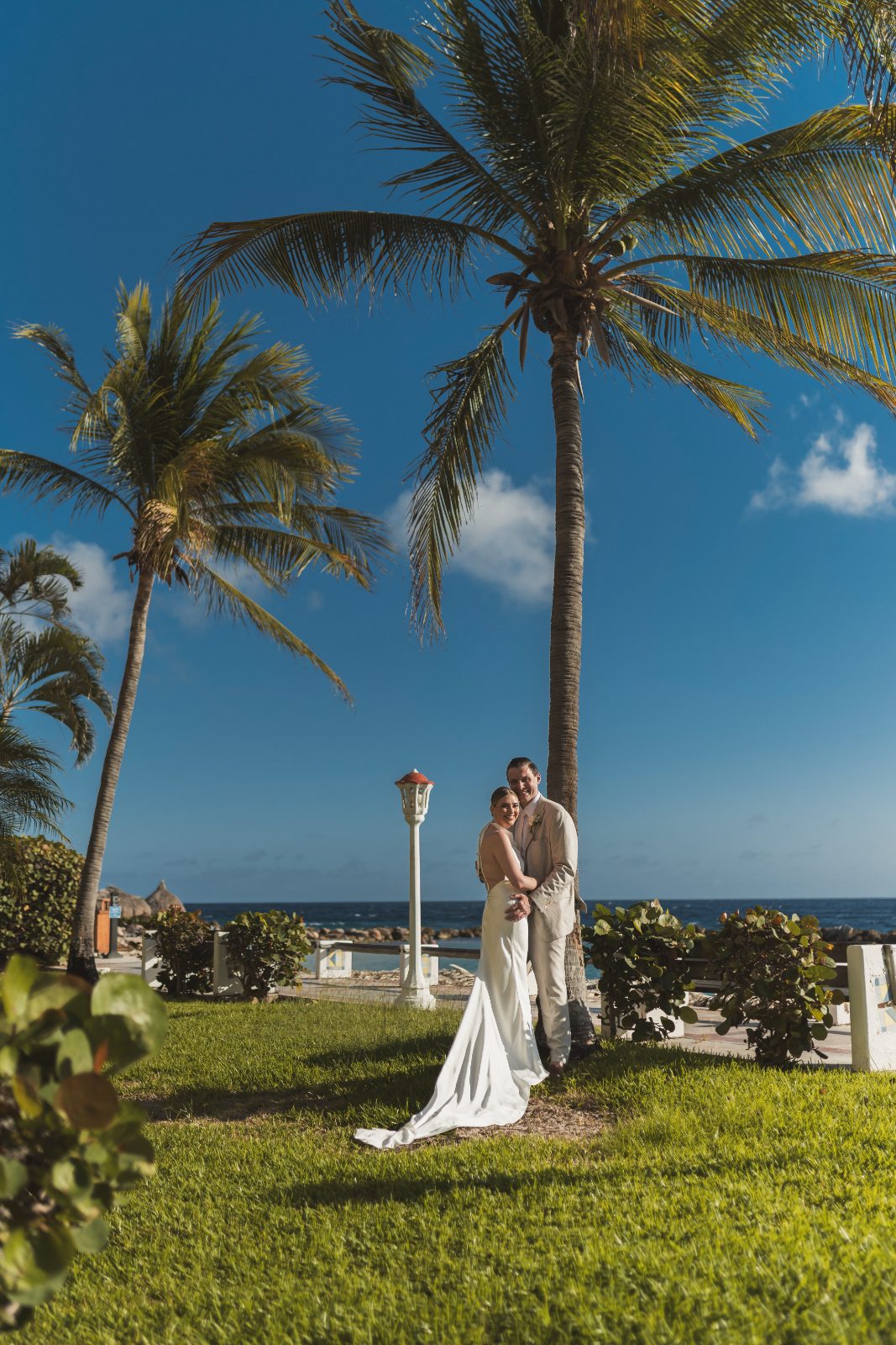 Jessica and Bogdan stand together in wedding attire at Avila Beach Hotel, surrounded by palm trees and a clear blue sky by the ocean