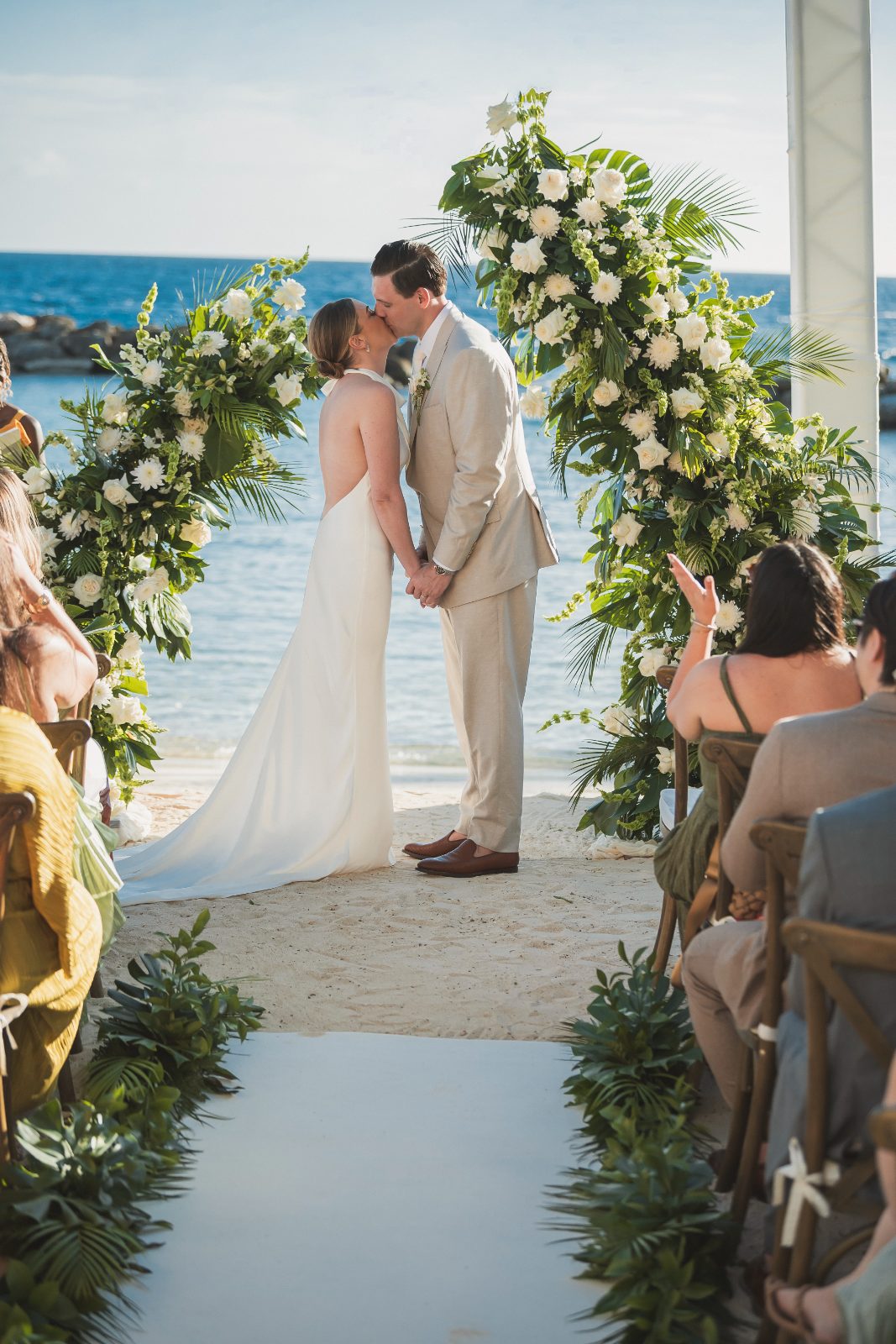 Jessica and Bogdan are kissing under an arch decorated with flowers at their wedding ceremony on the beach in Curaçao