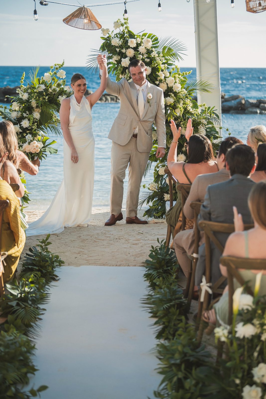 Jessica and Bogdan are celebrating their wedding at Avila Beach Hotel, Curaçao, with guests applauding as they raise their hands in front of floral decorations