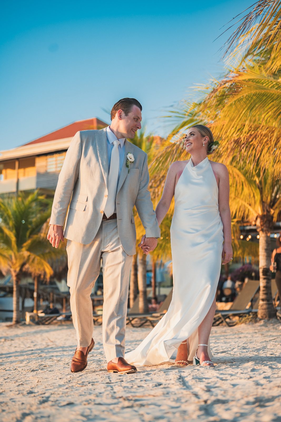 Jessica and Bogdan walk hand in hand on the beach at Avila Beach Hotel, Curaçao, surrounded by palm trees and a serene atmosphere