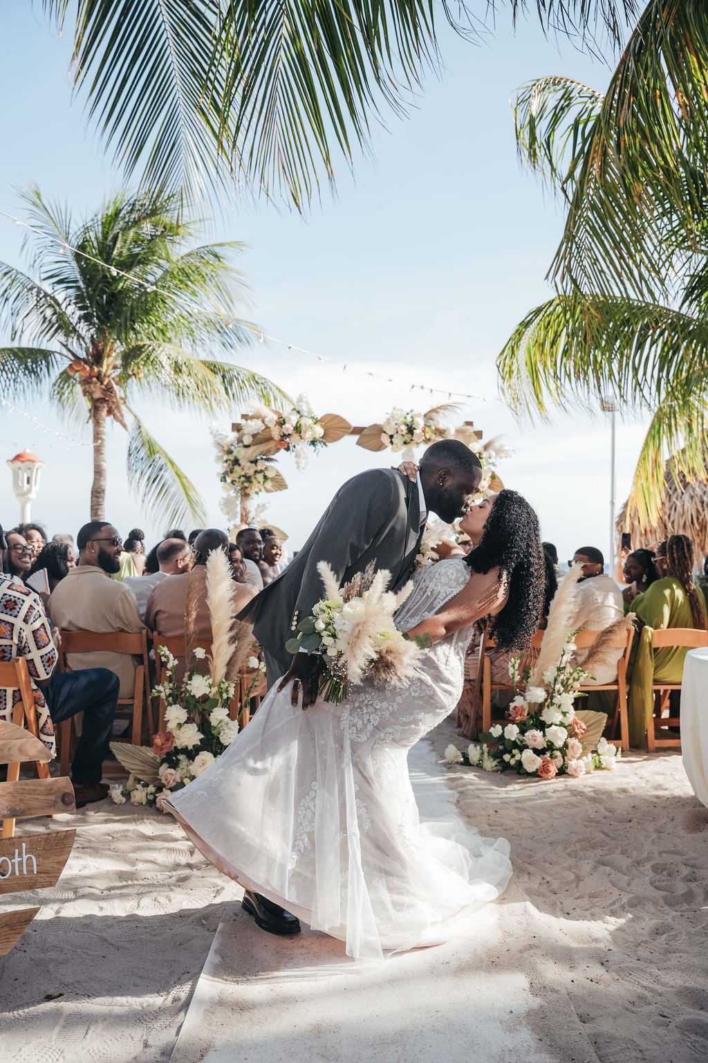 Kayla and Anthony kiss during their beach wedding ceremony at Avila Beach Hotel, with guests seated in the background and tropical decorations