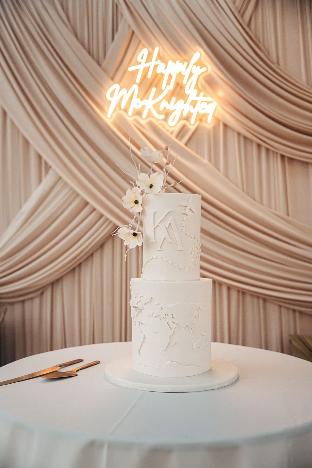 Kayla and Anthony's wedding cake is displayed at the Avila Beach Hotel, featuring white frosting and floral decoration, under a neon sign