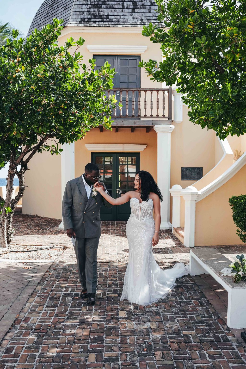 Kayla and Anthony walk hand in hand on a brick path at the Avila Beach Hotel, surrounded by greenery and a yellow building