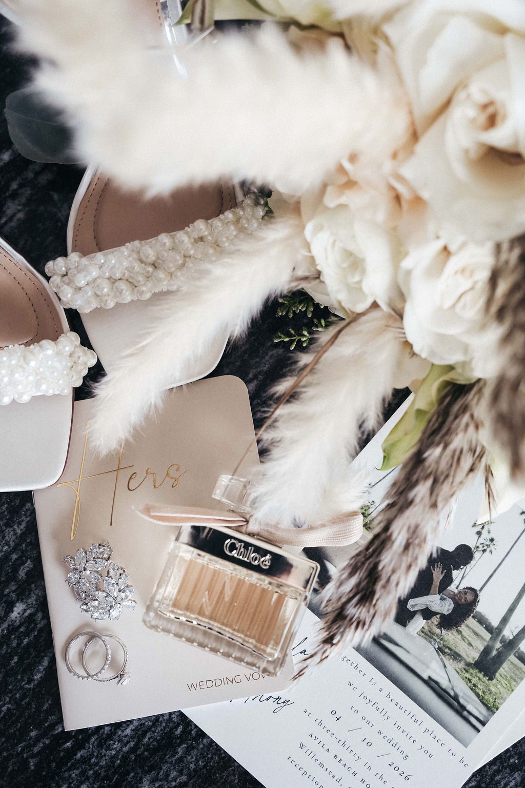 The scene shows wedding details, including shoes, perfume, floral bouquet, and invitations, set on a dark surface at Avila Beach Hotel