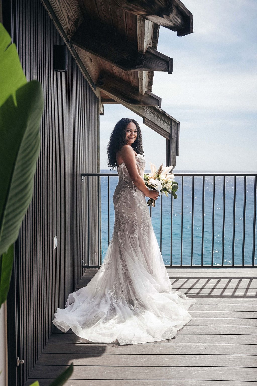 Kayla stands on a balcony at Avila Beach Hotel, wearing a wedding dress and holding a bouquet, with the ocean in the background