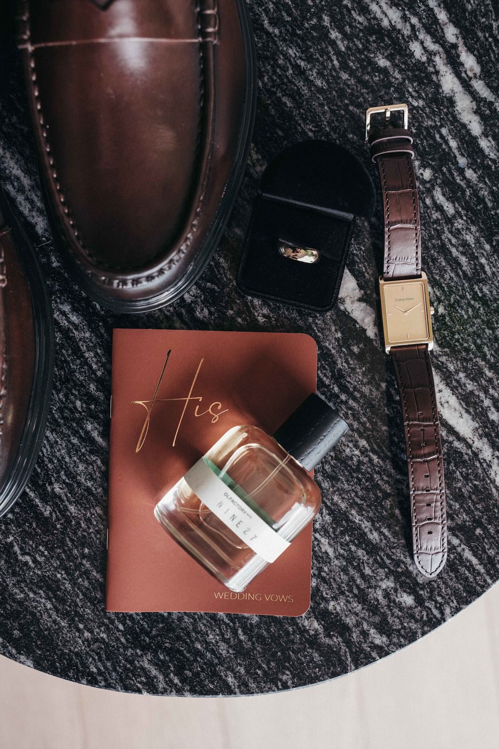 The wedding items, including shoes, a ring box, a watch, a perfume bottle, and vow book, are arranged on a dark marble surface