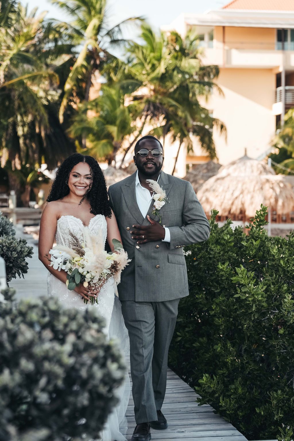 Kayla and Anthony walk side by side on a wooden pathway surrounded by greenery at the Avila Beach Hotel for their wedding